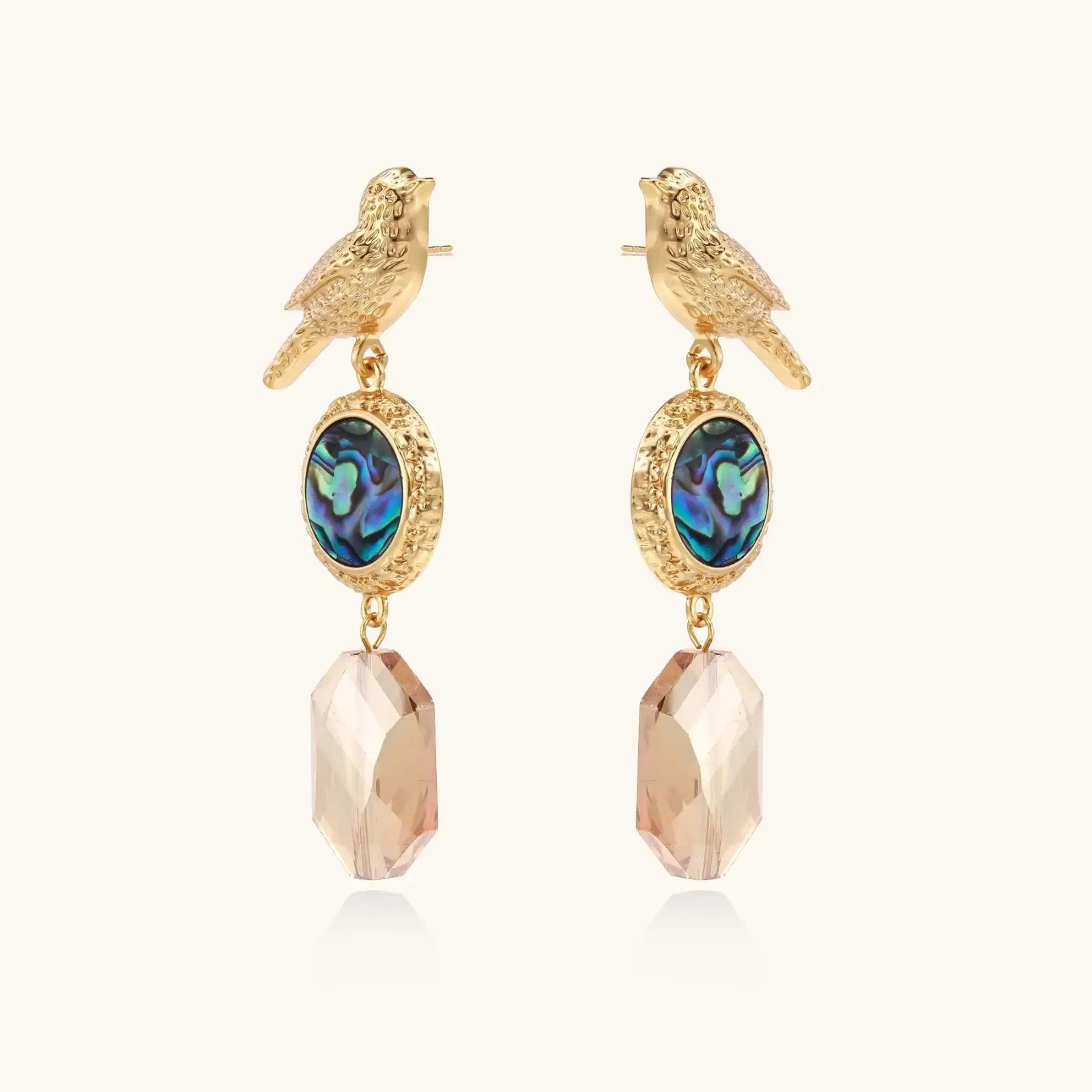 Gold-plated bird stud earrings with iridescent abalone shell medallions and champagne crystal drops on cream background